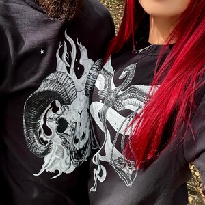 Op de afbeelding: Twee zwarte shirts met lange mouwen en witte grafische prints. Het ene shirt toont een gedetailleerde ramsschedel omringd door vlammen en kleine sterren. Het andere shirt toont een gevleugelde zwaan- of feniksfiguur die een weegschaal met een schedel vasthoudt.