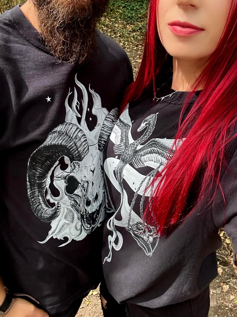 Op de afbeelding: Twee zwarte shirts met lange mouwen en witte grafische prints. Het ene shirt toont een gedetailleerde ramsschedel omringd door vlammen en kleine sterren. Het andere shirt toont een gevleugelde zwaan- of feniksfiguur die een weegschaal met een schedel vasthoudt.