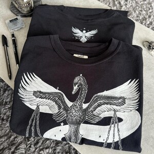 Op de afbeelding: Twee zwarte sweaters met ronde hals en een witte grafische print. Het ontwerp aan de voorzijde toont een grote zwaan met gespreide vleugels die kettingen in zijn klauwen houdt, omringd door kleine sterren. Een kleinere versie staat op de borst.