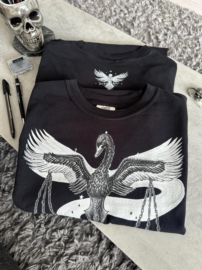 Op de afbeelding: Twee zwarte sweaters met ronde hals en een witte grafische print. Het ontwerp aan de voorzijde toont een grote zwaan met gespreide vleugels die kettingen in zijn klauwen houdt, omringd door kleine sterren. Een kleinere versie staat op de borst.