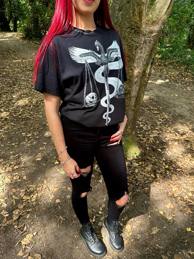 Op de afbeelding: Een zwart T-shirt met korte mouwen en een witte grafische print van een gevleugeld wezen dat lijkt op een zwaan of feniks, verweven met een kronkelend pad en een weegschaal met een schedel aan de ene kant en kristallen aan de andere.
