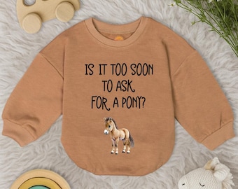 May include: A tan baby bodysuit with long sleeves. The text "IS IT TOO SOON TO ASK FOR A PONY?" is printed on the front, along with a graphic of a pony. The bodysuit is surrounded by wooden toys and a stuffed animal.