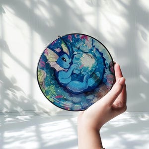 Może przedstawiać: Okrągłe witrażowe dzieło sztuki przedstawiające niebieską postać z kreskówki w wodnym otoczeniu. Dzieło trzymane jest w dłoni, podkreślając żywe kolory i szczegółowy design. Posiada czarną metalową ramę.