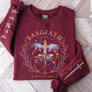 Peut inclure: Sweat-shirt bordeaux avec un motif graphique repr&eacute;sentant une &eacute;p&eacute;e, un dragon et le texte "BASGIATH WAR COLLEGE". Les manches comportent des illustrations suppl&eacute;mentaires et les mots "Violent little thing".
