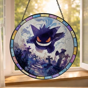 Może przedstawiać: Okrągłe witrażowe okno z fioletową postacią Gengara z pomarańczowym uśmiechem, osadzone na tle sceny cmentarnej. Okno ma czarną obwódkę i kolorową mozaikową krawędź, zawieszone na łańcuchu.