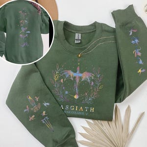 May include: A dark green sweatshirt with a crew neck and long sleeves. The front features a colorful graphic of a bird-like creature and the text "ASGIATH WAR COLLEGE". The sleeves and back have matching designs. A gold necklace is draped on the sweatshirt.