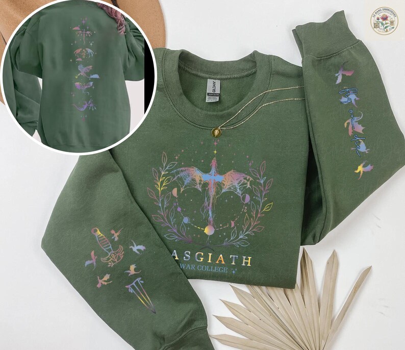 May include: A dark green sweatshirt with a crew neck and long sleeves. The front features a colorful graphic of a bird-like creature and the text "ASGIATH WAR COLLEGE". The sleeves and back have matching designs. A gold necklace is draped on the sweatshirt.