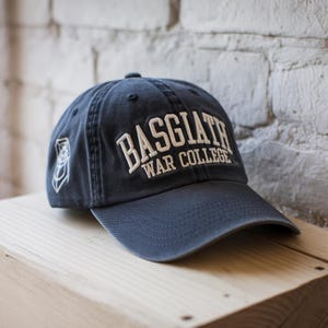 以下が含まれることがあります： 「BASGIATH WAR COLLEGE」の文字が白で刺繍された、ネイビーブルーの野球帽。つばはカーブしており、側面に小さなエンブレムが付いています。帽子は木製の表面に置かれています。