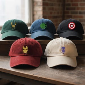 Può includere: Cinque cappellini da baseball in vari colori, ognuno con un diverso emblema di supereroe. I cappellini sono verde scuro, blu navy, nero, bordeaux e beige. Gli emblemi includono un elmo dorato, un pugno verde, uno scudo rosso e bianco, una maschera dorata e una maschera viola.