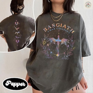 Puede incluir: Camiseta gris oscuro con un gráfico de dragón colorido y el texto "BASGIATH WAR COLLEGE". La parte trasera de la camiseta presenta un diseño similar con dragones estilizados. El diseño incluye elementos celestiales y una corona floral.