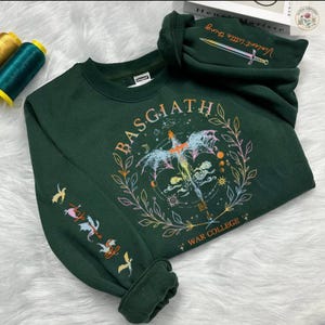 Peut inclure: Sweat-shirt vert fonc&eacute; avec le mot "BASGIATH" dans un motif circulaire, repr&eacute;sentant une cr&eacute;ature ail&eacute;e et les mots "WAR COLLEGE". La manche pr&eacute;sente des illustrations color&eacute;es d'oiseaux et d'autres symboles. La capuche affiche le texte "Violent little thing" et une &eacute;p&eacute;e.