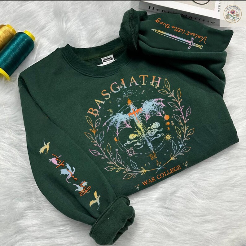 Peut inclure: Sweat-shirt vert fonc&eacute; avec le mot "BASGIATH" dans un motif circulaire, repr&eacute;sentant une cr&eacute;ature ail&eacute;e et les mots "WAR COLLEGE". La manche pr&eacute;sente des illustrations color&eacute;es d'oiseaux et d'autres symboles. La capuche affiche le texte "Violent little thing" et une &eacute;p&eacute;e.