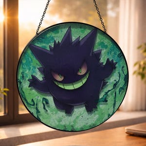 Może przedstawiać: Okrągłe witrażowe dzieło sztuki przedstawiające fioletową postać z kreskówki z szerokim uśmiechem i zielonymi oczami. Postać jest osadzona na zielono-niebieskim tle. Grafika jest oprawiona w czarny metalowy okrąg i zawieszona na łańcuchu.