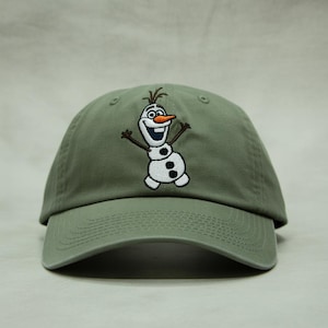 May include: Olive green baseball cap featuring an embroidered image of Olaf, the snowman character from the movie Frozen. The cap has a curved brim and an adjustable strap at the back. The Olaf design includes a carrot nose, twig arms, and a smiling face.