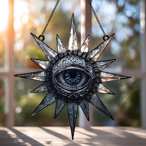 May include: A star-shaped wall hanging with a central eye design. The eye has a blue iris and is surrounded by ornate detailing. The star points are silver and the piece is suspended by a chain. The background is blurred, suggesting a window.