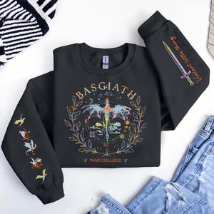 Peut inclure: Sweat-shirt noir avec un motif graphique color&eacute; repr&eacute;sentant un dragon, une &eacute;p&eacute;e et le texte "BASGIATH WAR COLLEGE". Les manches affichent des illustrations suppl&eacute;mentaires et les mots "Violent Little Thing".