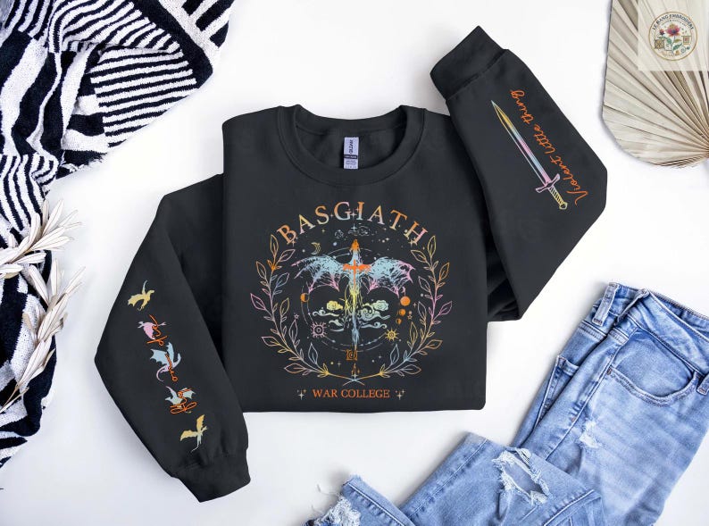 Peut inclure: Sweat-shirt noir avec un motif graphique color&eacute; repr&eacute;sentant un dragon, une &eacute;p&eacute;e et le texte "BASGIATH WAR COLLEGE". Les manches affichent des illustrations suppl&eacute;mentaires et les mots "Violent Little Thing".
