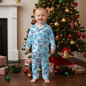 Puede incluir: Un ni&ntilde;o lleva un pijama azul claro con un estampado repetido de personajes de dibujos animados y Pokeballs. El pijama incluye una camisa con botones y pantalones a juego. El ni&ntilde;o est&aacute; de pie frente a un &aacute;rbol de Navidad decorado.