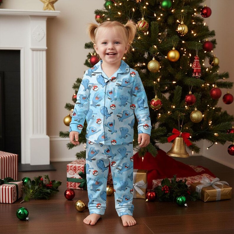 Puede incluir: Un ni&ntilde;o lleva un pijama azul claro con un estampado repetido de personajes de dibujos animados y Pokeballs. El pijama incluye una camisa con botones y pantalones a juego. El ni&ntilde;o est&aacute; de pie frente a un &aacute;rbol de Navidad decorado.