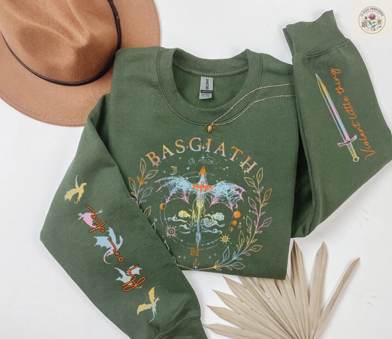 Peut inclure: Un sweat-shirt vert olive avec le mot "BASGIATH" dans un motif circulaire avec une &eacute;p&eacute;e et des &eacute;l&eacute;ments floraux. La manche droite porte le texte "Violent little thing" et un dessin d'&eacute;p&eacute;e. La manche gauche a des graphiques de dragons.