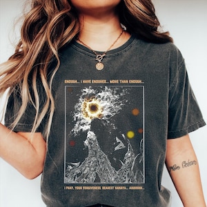 以下が含まれることがあります： 明るい太陽のような画像と人物が描かれたグラフィックデザインのダークグレーのTシャツ。「ENOUGH... I HAVE ENDURED... MORE THAN ENOUGH... I PRAY, YOUR FORGIVENESS, DEAREST NANAYA... AHHHHHH...」と書かれたテキスト。
