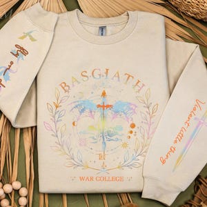 Peut inclure: Sweat-shirt beige avec un motif graphique color&eacute; repr&eacute;sentant une &eacute;p&eacute;e, des &eacute;l&eacute;ments c&eacute;lestes et le texte "BASGIATH WAR COLLEGE". Les manches affichent les phrases "Fly me die" et "Violent little thing."