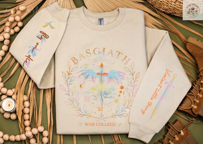 Peut inclure: Sweat-shirt beige avec un motif graphique color&eacute; repr&eacute;sentant une &eacute;p&eacute;e, des &eacute;l&eacute;ments c&eacute;lestes et le texte "BASGIATH WAR COLLEGE". Les manches affichent les phrases "Fly me die" et "Violent little thing."