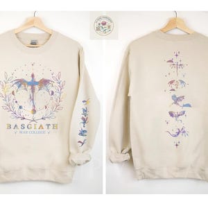 May include: Beige sweatshirt with a dragon graphic on the front and back. The front design features a dragon with a colorful wing design, surrounded by a wreath and the text "BASGIATH WAR COLLEGE". The sleeves have additional dragon illustrations.
