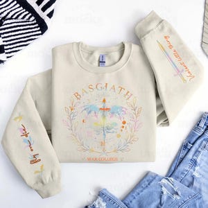 Peut inclure: Sweat-shirt cr&egrave;me avec le mot "BASGIATH" dans un motif circulaire, avec une &eacute;p&eacute;e et des &eacute;l&eacute;ments floraux. Les manches ont du texte et un motif d'&eacute;p&eacute;e. Une paire de jeans bleus est visible.