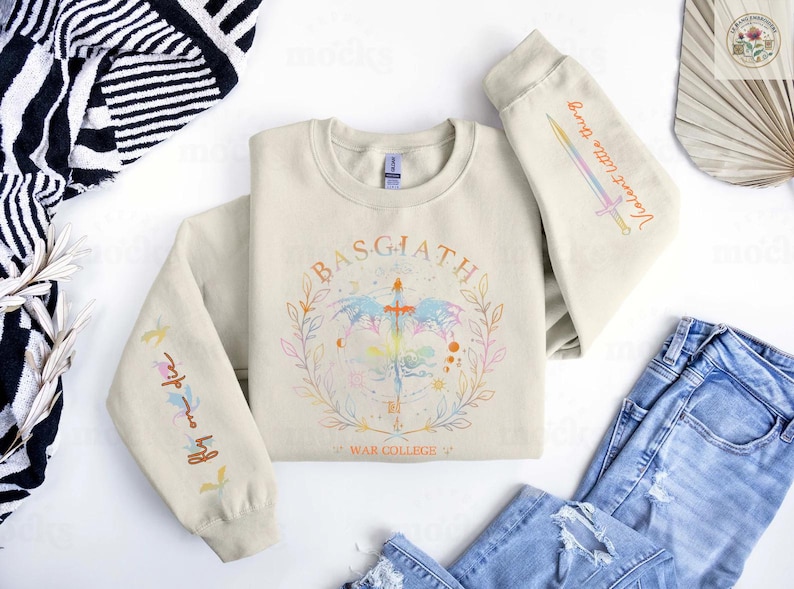 Peut inclure: Sweat-shirt cr&egrave;me avec le mot "BASGIATH" dans un motif circulaire, avec une &eacute;p&eacute;e et des &eacute;l&eacute;ments floraux. Les manches ont du texte et un motif d'&eacute;p&eacute;e. Une paire de jeans bleus est visible.