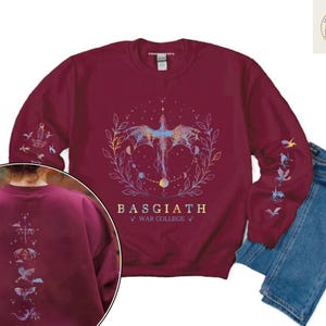 May include: Burgundy sweatshirt with a celestial dragon design. The design includes a dragon, moons, and stars, with the text "BASGIATH WAR COLLEGE". Additional dragon and celestial designs are on the sleeves and back. Blue jeans are next to the sweatshirt.