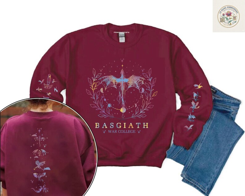 May include: Burgundy sweatshirt with a celestial dragon design. The design includes a dragon, moons, and stars, with the text "BASGIATH WAR COLLEGE". Additional dragon and celestial designs are on the sleeves and back. Blue jeans are next to the sweatshirt.