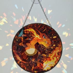Może przedstawiać: Okrągły witrażowy łapacz słońca z motywem Charizarda. Grafika przedstawia ognisty Pokémon w odcieniach pomarańczu, czerwieni i żółci, z płomieniami i ciemnym tłem. Wisi na łańcuszku i przyssawce, idealny do okna.