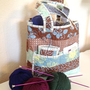 May include: A brown and blue patchwork tote bag with multiple pockets. The bag is filled with balls of yarn and knitting needles.