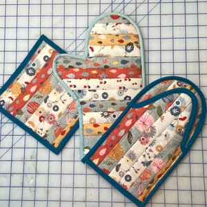 May include: A quilted oven mitt and two square pot holders featuring a horizontal striped pattern of floral and leaf motifs in shades of red, orange, yellow, blue, and grey, all finished with solid blue fabric binding along the edges.