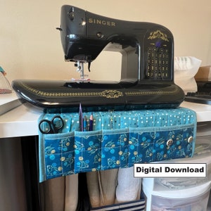 May include: A blue and white quilted sewing machine organizer with pockets for storing sewing supplies. The organizer is hanging on a white table with a black Singer sewing machine on top.
