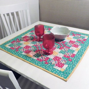 May include: A square, quilted table topper with a patchwork design in pink, white, and yellow squares, bordered by a teal fabric with white polka dots. Two red wine glasses and a white bowl sit on the table.