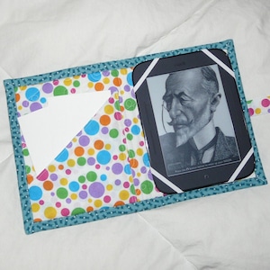 May include: A teal blue fabric e-reader case with a polka dot pattern in pink, yellow, green, blue, and purple. The case has a flap that folds over the e-reader and a pocket on the front. The e-reader is a Kindle with a black and white screen showing a man with a beard and glasses. The text on the screen reads "Press the 'P' button below to wake up your Kindle."