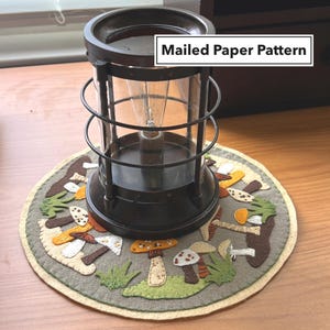 May include: A brown and black metal candle warmer with a clear glass shade sits on a round, gray felt rug with an embroidered mushroom design. The rug has a beige border. The text "Mailed Paper Pattern" is visible in the upper right corner of the image.