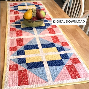 May include: A yellow, blue, red and pink quilted table runner with a geometric pattern. The table runner is on a wooden table with a bowl of fruit.