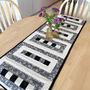 May include: A rectangular table runner featuring a geometric pattern of black, white, and gray fabric strips. The design includes alternating blocks of solid black squares and white fabric with a small gray floral print. Two clear glass vases hold purple flowers.