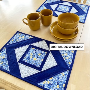 May include: Blue and white floral fabric placemat with a navy blue border and a geometric pattern. The placemat is on a wooden table with two yellow ceramic mugs and a yellow ceramic bowl.
