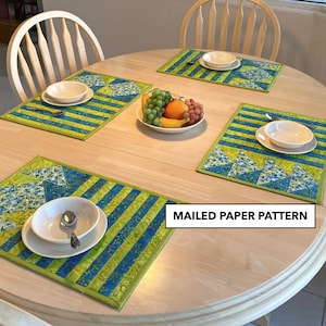 May include: A round wooden table set with four placemats, each with a blue and green striped pattern and a geometric design. White bowls and spoons are on the placemats. A bowl of fruit sits in the center. The text "MAILED PAPER PATTERN" is in a white box.