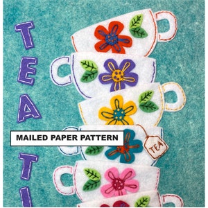 May include: A stack of five felt teacups with embroidered flowers in different colors. The teacups are white with a white outline. The teacups are stacked on a turquoise background. The text "MAILED PAPER PATTERN" is at the bottom of the image. A small felt tea bag with the word "TEA" is attached to the top teacup.