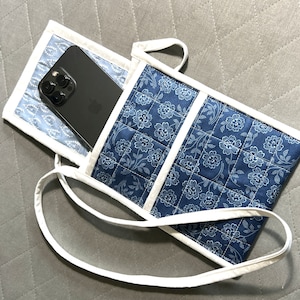 May include: A quilted phone bag with a floral pattern in shades of blue and white. The bag has a white strap and trim, and a black phone is partially visible inside. The bag is designed to hold a phone.