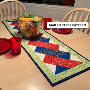 May include: A table runner with a chevron pattern in red, white, blue, and green fabrics. The runner is on a wooden table with a glass bowl of fruit and two red glasses.