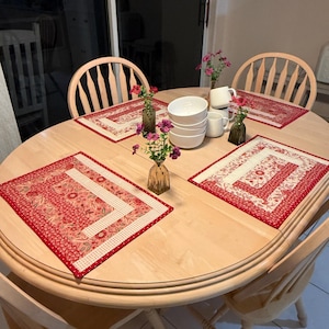 May include: A light wood oval dining table set with four rectangular fabric placemats featuring a red and white floral pattern. The table includes a stack of white ceramic bowls, two white mugs, and three small glass vases with pink flowers.