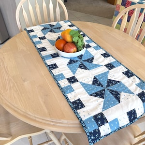 Puede incluir: Un camino de mesa acolchado rectangular con un patrón repetitivo de molinillos en tonos azul marino, azul claro y blanco. Un cuenco blanco se sitúa en el centro, lleno de una pera amarilla, una manzana roja, una naranja y uvas verdes.