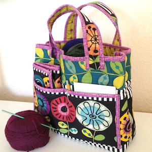 May include: A black and white tote bag with colorful floral patterns and purple trim. The bag has multiple pockets and is perfect for carrying knitting supplies.