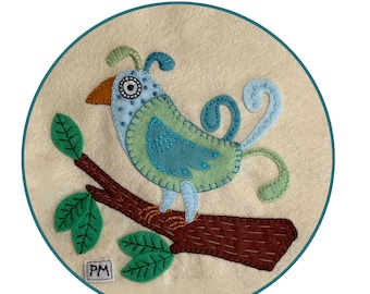 May include: A whimsical felt appliqué of a blue and green bird perched on a brown branch with green leaves. The bird has a large eye and a long, curly tail. The artwork is signed "PM" in the lower left corner.